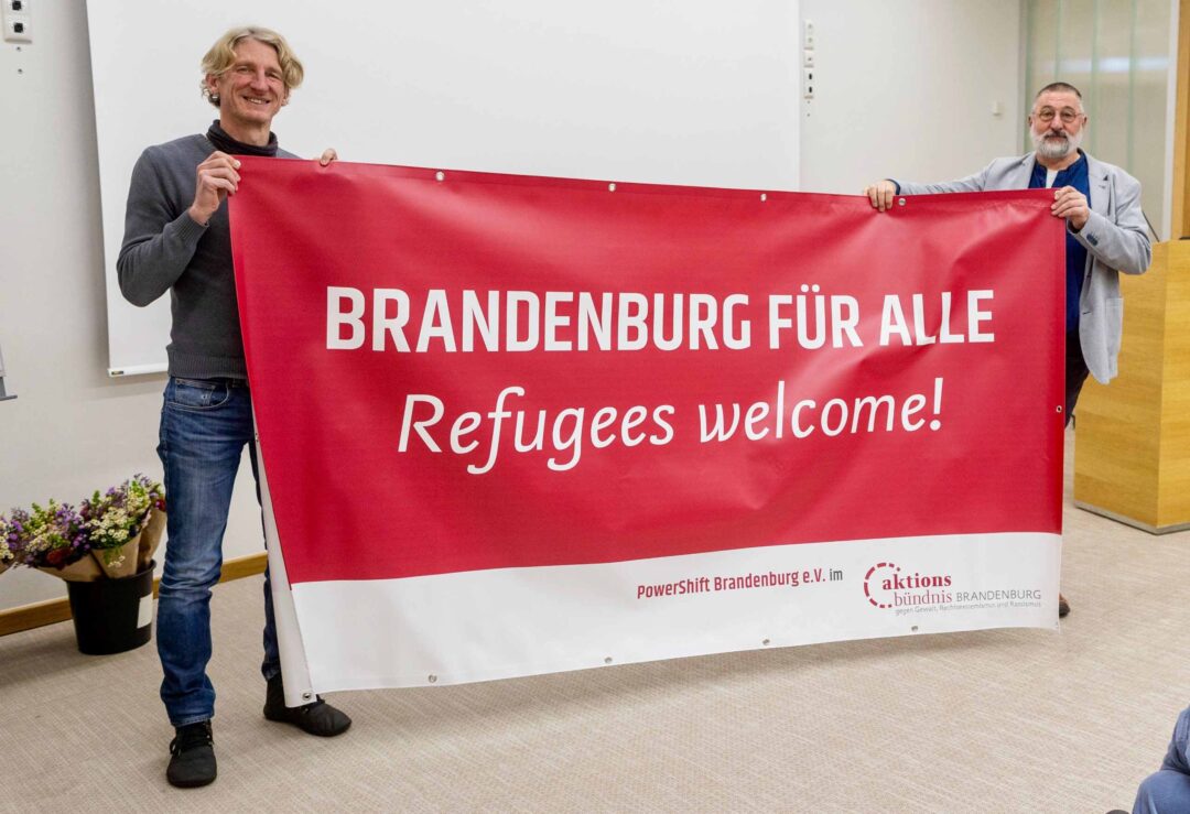 Thomas Wisch, Vorsitzender des Aktionsbündnis Brandenburg, und Bernd Brouns von PowerShift Brandenburg halten ein Banner mit der Aufschrift 'Brandenburg für Alle. Refugees welcome!'.